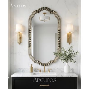 May include: An oval mirror with a decorative black and white border is the focal point of this bathroom scene. Gold sconces flank the mirror, and a black vanity with gold hardware sits below. The brand name "Arcuros" is displayed at the top and bottom of the image.