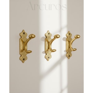 May include: Three ornate brass wall hooks with a decorative design. Each hook has a curved double hook design and is mounted on a rectangular backplate. The word "Arcuros" is printed at the top of the image.