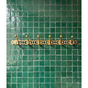 Puede incluir: Un perchero de pared de latón con seis ganchos redondeados. El perchero presenta un diseño decorativo con patrones geométricos verdes, blancos y negros. El perchero está montado en una pared de azulejos verdes.