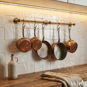 Könnte beinhalten: Eine Küchenszene mit einem Topfgestell aus Messing, an dem fünf Kupferpfannen hängen. Eine Keramikflasche mit Korken und ein Leinentuch liegen auf einer Holztheke vor einer weiß gefliesten Wand.