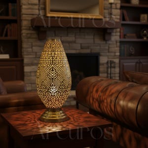 May include: A decorative table lamp with a gold-colored, patterned metal shade. The lamp has a teardrop shape and sits on a round base. The lamp is on a dark brown wooden table. The lamp is in a living room setting.