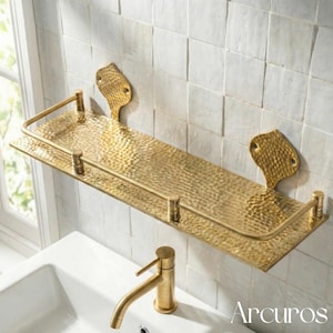 May include: A hammered brass bathroom shelf with a curved front rail and decorative wall mounts. The shelf is mounted above a white sink and a brass faucet. The word "Arcuros" is visible in the bottom right corner.