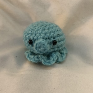 May include: A small, handmade crochet octopus in a light blue color. The octopus has a rounded head, a small, round body, and eight curled tentacles. It has two small, black eyes. The octopus is made of soft yarn.
