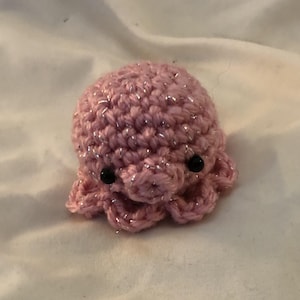 May include: A small, handmade crochet octopus in a soft pink color with silver metallic thread accents. The octopus has black bead eyes and eight curled tentacles. This plush toy is a cute and whimsical item.