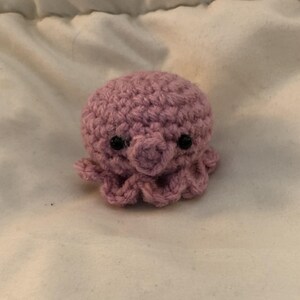 May include: A small, handmade crochet octopus in a soft mauve color. The octopus has a rounded head, small tentacles, and two black safety eyes. The plush toy is made of a textured yarn.
