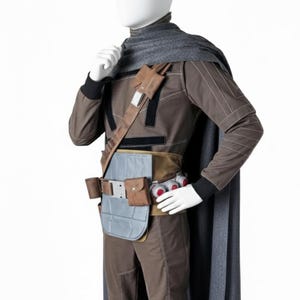 May include: A brown and gray costume with a cape, belt, and accessories. The outfit includes a long-sleeved top, pants, and a gray cape. The belt has pouches and a gray chest plate. The costume is displayed on a mannequin.