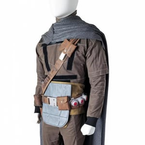 May include: A brown and gray costume with a cape, designed to resemble a character from a science fiction film. The outfit includes a jacket, pants, belt, and accessories. The mannequin is wearing a gray scarf and a brown shoulder strap.