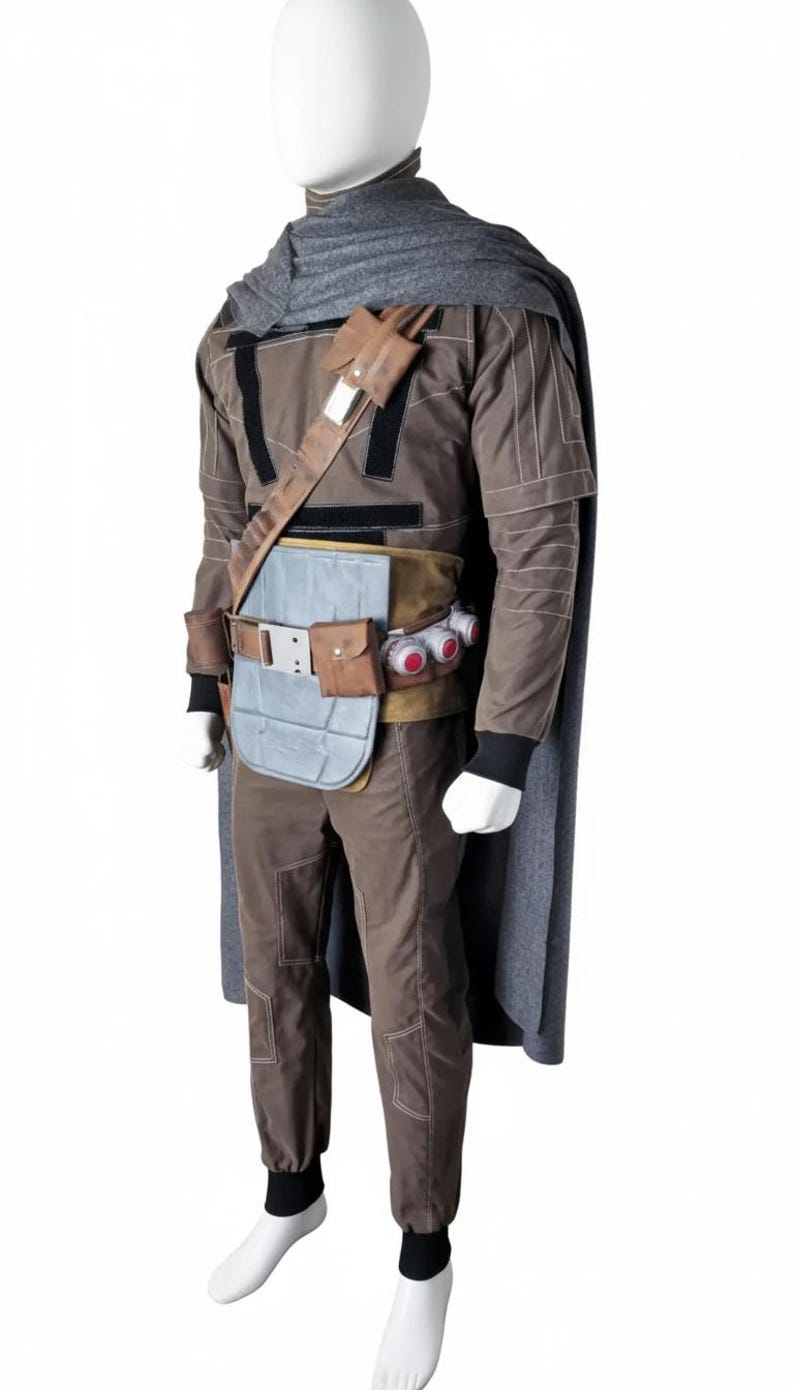 May include: A brown and gray costume with a cape, designed to resemble a character from a science fiction film. The outfit includes a jacket, pants, belt, and accessories. The mannequin is wearing a gray scarf and a brown shoulder strap.