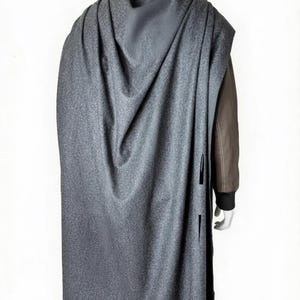May include: A gray cape with a textured pattern, draped over a mannequin. The cape has a long, flowing design and features vertical slits on the side. The mannequin is wearing a brown jacket underneath.