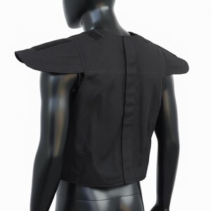 May include: Black tactical shoulder and upper chest protector. The protective gear has a padded design with wide shoulder flaps and a central vertical seam. The item is displayed on a black mannequin against a white background.