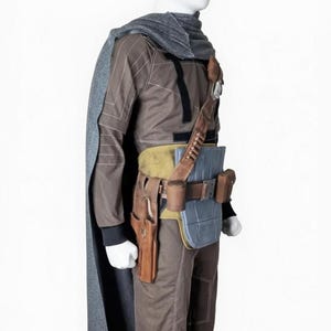 May include: A brown and gray costume with a cape, designed to resemble a character from a science fiction film. The outfit includes a long-sleeved top, pants, and a utility belt with pouches and a holster. A gray scarf and cape complete the ensemble.
