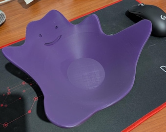 May include: A purple, 3D-printed Ditto-shaped bowl with a smiling face. The bowl is made of a smooth, solid material and has a circular indentation in the center. The bowl is sitting on a black and red mousepad.