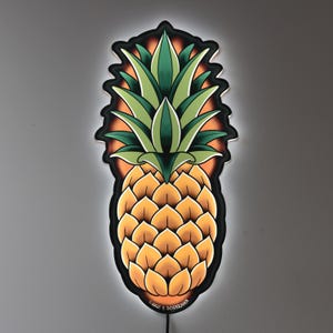 May include: A decorative pineapple-shaped wall art piece with a black outline and a glowing effect. The pineapple features a detailed design with green leaves and a yellow-orange body. The artwork is mounted on a gray wall.