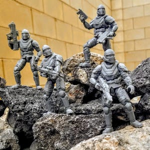 May include: Four gray action figures of soldiers in combat gear, posed on a rocky surface. Each figure is armed with a weapon and wears a helmet. The background is a textured, light-colored wall.
