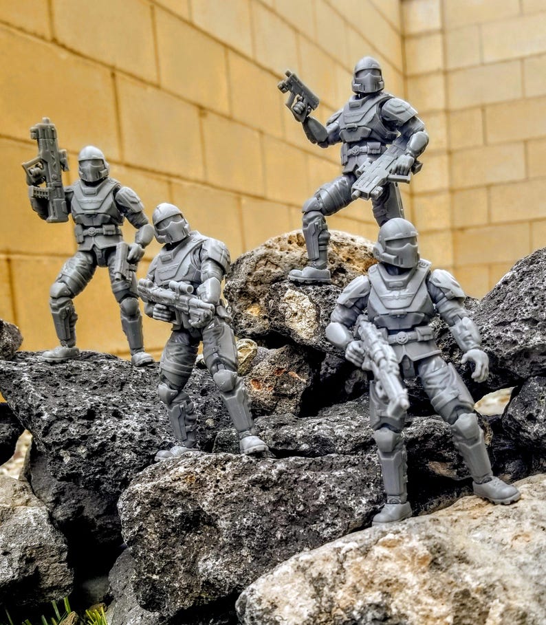 May include: Four gray action figures of soldiers in combat gear, posed on a rocky surface. Each figure is armed with a weapon and wears a helmet. The background is a textured, light-colored wall.
