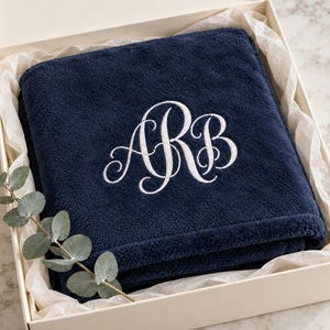 May include: A folded navy blue plush blanket featuring white embroidered monogram letters ARB in a cursive script font. The blanket is presented inside a white gift box lined with white tissue paper and decorated with a sprig of green eucalyptus.