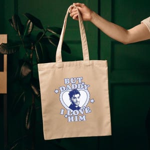Puede incluir: Bolso tote beige con el texto "BUT DADDY I LOVE HIM" en azul y blanco. El diseño incluye un gráfico en forma de corazón con un retrato de un hombre. El bolso es sostenido por una persona.