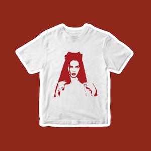 May include: White t-shirt featuring a red graphic of a woman with long hair and a bold makeup look. The design is a stylized, pop art-inspired illustration. The shirt is displayed on a red background.
