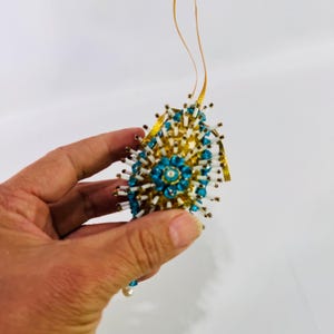 May include: A decorative egg-shaped ornament featuring a gold-toned base adorned with numerous white cylindrical beads and bright blue faceted crystals arranged in a floral pattern, suspended by a thin gold ribbon loop for hanging.