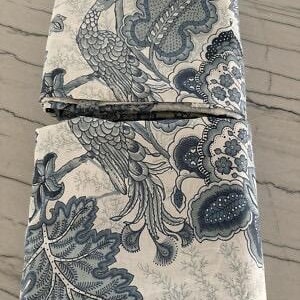 May include: Two folded fabric pieces with a blue and white peacock and floral pattern. The design features a peacock perched on a branch, surrounded by large leaves and floral elements. The fabric has a detailed, intricate design.