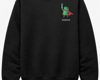 May include: Black sweatshirt featuring a green frog character wearing a red cape and holding a sword. The word "Huzzah" is printed below the frog. The sweatshirt is a crew neck style.