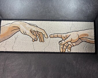 May include: A rectangular embroidered patch featuring a beige background with a black border. The design depicts two light-skinned hands reaching toward each other, with fingers nearly touching, rendered in shades of tan, peach, and brown thread.
