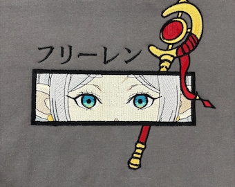 May include: A charcoal gray sweatshirt featuring an embroidered rectangular panel with a pair of bright blue eyes and white hair. Above the panel is the Japanese text フリーレン. A gold staff with a red gem and red ribbon extends from the panel.