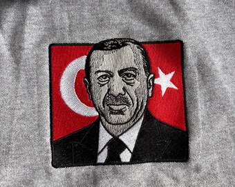 May include: Embroidered patch featuring a portrait of a man in a suit, set against a red background with a white crescent moon and star, resembling a flag. The patch is sewn onto a gray fabric.