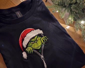 May include: Black t-shirt featuring an embroidered Grinch character wearing a Santa hat. The Grinch is emerging from a zipper design. The shirt is a festive holiday item.
