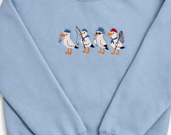 May include: Light blue sweatshirt featuring an embroidered design of four seagulls, each with fishing-related accessories. The seagulls are wearing hats, sunglasses, and carrying fishing gear. The sweatshirt has a crew neck and long sleeves.
