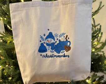 May include: A white canvas tote bag with a festive winter design. The design features a snowman, snowflakes, a mug of hot chocolate, and the text "#christmasvibes" in blue. The bag has white handles.