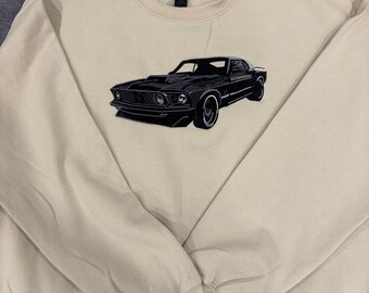 May include: A beige sweatshirt featuring a black embroidered classic muscle car design. The car is detailed with a hood scoop and racing stripes. The sweatshirt has a crew neck and long sleeves.