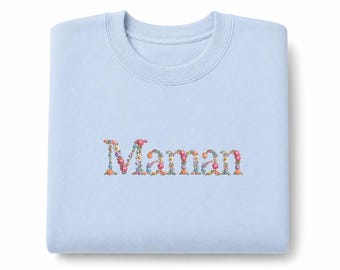 May include: A light blue sweatshirt with the word "Maman" embroidered in colorful floral letters. The sweatshirt is folded neatly, showcasing the floral embroidery in shades of pink, yellow, blue, and green.