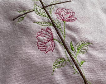 May include: A light pink sweater with an embroidered design. The design features a brown cross with a stem and two pink roses with green leaves. The embroidery is detailed and adds a decorative element to the sweater.
