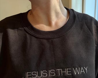 May include: Black crewneck sweatshirt with the embroidered text "JESUS IS THE WAY" in white. The sweatshirt is a solid black color and has a classic, casual design. The text is centered on the chest.