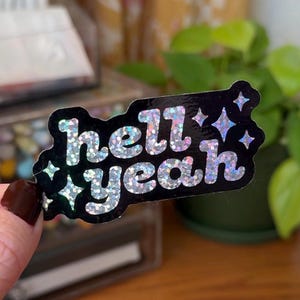 May include: A black sticker with the words "hell yeah" in silver glitter lettering. The sticker has a black border with silver glitter star accents. The sticker is held by a person with dark nail polish.