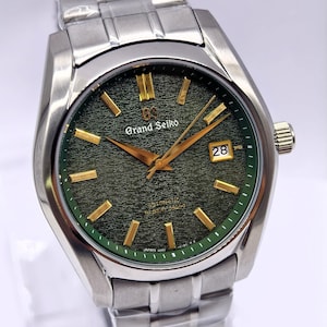 Op de afbeelding: Een Grand Seiko polshorloge van roestvrij staal met een groene wijzerplaat en goudkleurige wijzers en markeringen. Het horloge heeft een datumweergave op 3 uur en een roestvrijstalen band. Een luxe item.