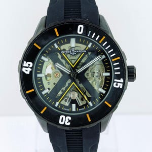 May include: Black Ulysse Nardin watch with a black rubber strap. The watch face has a skeleton design with visible gears and a black and yellow colour scheme. The bezel features white and orange markings.