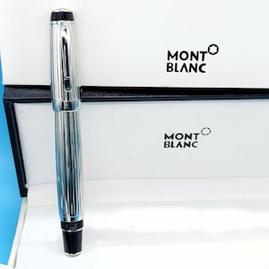 Könnte beinhalten: Ein silberner Montblanc-Stift mit schwarzer Spitze und Kappe, präsentiert in einer weißen Schachtel mit dem Montblanc-Logo. Der Stift hat ein geripptes Design. Die Schachtel ist schwarz und weiß.