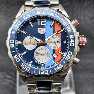 Könnte beinhalten: Eine TAG Heuer Formula 1 Chronographenuhr aus Edelstahl mit blauem Zifferblatt und einem vertikalen hellblauen und orangefarbenen Rennstreifen. Die Uhr verfügt über drei silberne Hilfszifferblätter, eine Tachymeterlünette und ein Metallgliederarmband mit blauen Akzenten.