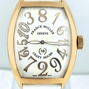 May include: A gold-toned Franck Muller Crazy Hours watch with a white dial and white leather strap. The watch face features large, stylized numerals and the text "CRAZY HOURS". The watch has a rectangular case and a textured crown.