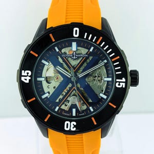 Pode incluir: Um relógio Ulysse Nardin preto com uma bracelete de borracha laranja. O mostrador do relógio tem um design de esqueleto com detalhes em azul e laranja. A luneta apresenta algarismos brancos e marcadores laranja.