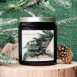 May include: A black glass candle jar with a gold lid, labeled "Irch Fraser Fir." The label features a green fir branch illustration and text. A pine cone rests beside the candle on a wooden surface, with a backdrop of evergreen branches.