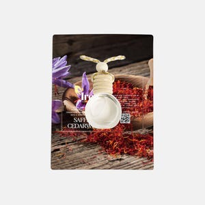 May include: A clear, round air freshener with a wooden top and a decorative butterfly design. The air freshener is labeled "Saffron Cedarwood" and is displayed with saffron threads and purple flowers.