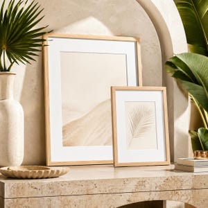 May include: Two framed art prints with light wood frames and white mats. One print features a beige landscape, the other a beige palm leaf. The frames are displayed on a stone surface, with a vase and decorative bowl in the foreground.