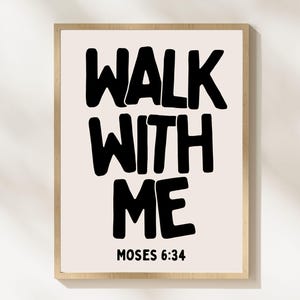 May include: A framed print with the words "WALK WITH ME" in bold black letters on a cream background. Below the text is "MOSES 6:34" in smaller black letters. The frame is a light wood color.