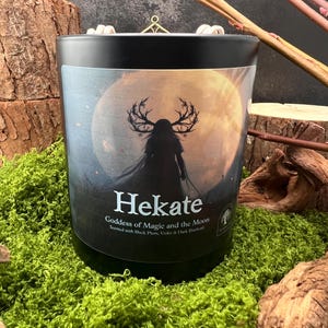 May include: A black candle with a label depicting a figure with antlers against a full moon. The label reads "Hekate Goddess of Magic and the Moon". The candle sits on green moss, with wood pieces in the background.