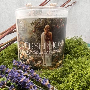 Puede incluir: Una vela cilíndrica con una etiqueta que presenta una ilustración de una mujer con un vestido blanco, con las palabras "PERSEPHONE Goddess of Spring". La vela está rodeada de musgo verde, lavanda morada y ramitas.