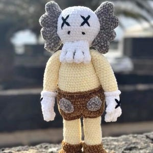 Puede incluir: Muñeca de ganchillo hecha a mano. La muñeca tiene una cabeza blanca con ojos de X negros, orejas grises, un cuerpo crema, pantalones cortos marrones, guantes blancos y zapatos marrones con detalles de X negros.