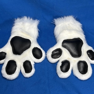 May include: A pair of white furry paw gloves with black paw pads and toe beans. The gloves are designed to resemble animal paws, with a fluffy texture and a realistic appearance. The gloves are set against a blue background.